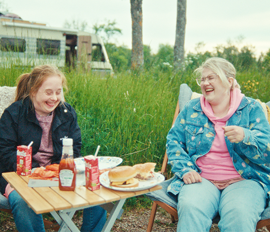 Bild från filmen "Det kunde varit vi" - Två glada tjejer sitter och skrattar tillsammans utomhus framför ett picnicbord med mat